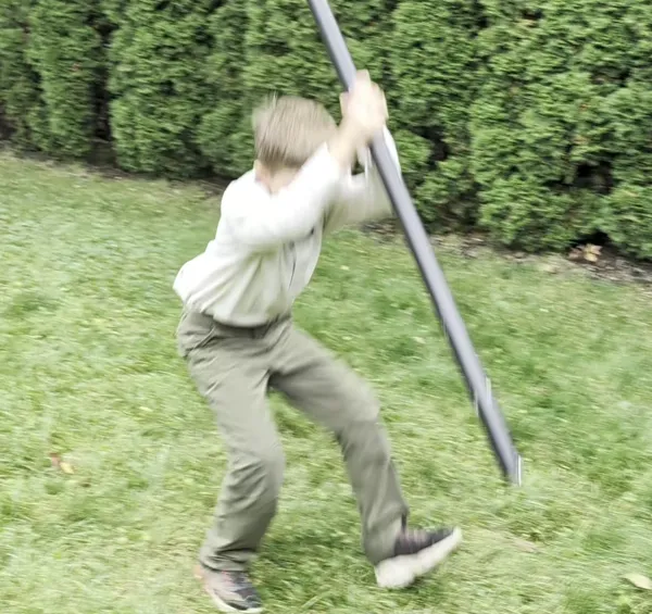 A boy swings a long pole down in a vertical strike on a lawn, seen in motion.